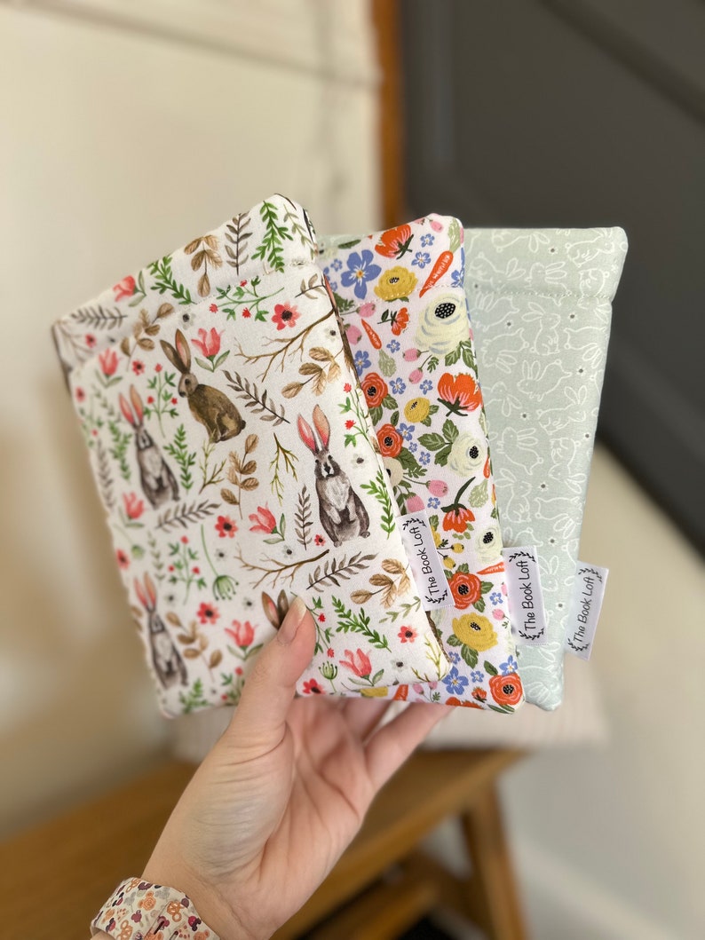 K&ouml;nnte beinhalten: Drei Stoffbeutel mit Blumen- und Hasenmustern. Die Beutel sind wei&szlig; mit verschiedenen farbigen Blumendesigns und haben ein wei&szlig;es Hasenmuster auf der R&uuml;ckseite. Jeder Beutel hat ein wei&szlig;es Etikett mit dem aufgedruckten Text "The Book Loft".