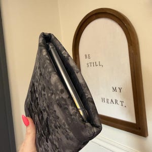 May include: A dark gray floral patterned phone sleeve with a phone inside. The sleeve is held in a hand with pink nail polish. A framed sign in the background reads "BE STILL, MY HEART."