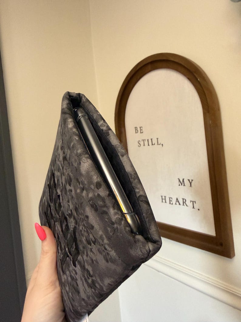 May include: A dark gray floral patterned phone sleeve with a phone inside. The sleeve is held in a hand with pink nail polish. A framed sign in the background reads "BE STILL, MY HEART."
