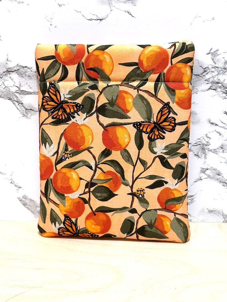 May include: A peach-coloured fabric pouch with a zipper closure. The pouch features a repeating pattern of orange citrus fruit, green leaves, and butterflies.
