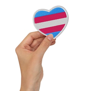 Pode incluir: Um patch bordado em forma de coração com as cores da bandeira do orgulho transgênero: azul claro, rosa e branco. O patch é segurado em uma mão contra um fundo branco. O patch tem aproximadamente 7.6 cm de largura.