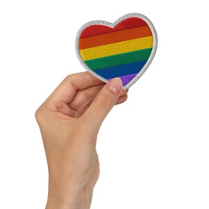 May include: Embroidered heart-shaped patch featuring a rainbow design with red, orange, yellow, green, blue, and purple stripes. The patch has a white border and is held against a white background.