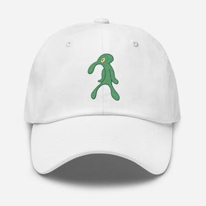 Peut inclure: Casquette de baseball blanche ornée d'un dessin de personnage de dessin animé vert. Le personnage est une silhouette verte stylisée avec une grosse tête et un seul œil. La casquette a une visière incurvée et une sangle réglable.