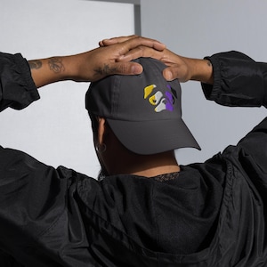 Könnte beinhalten: Dunkelgraue Baseballkappe mit gebogenem Schirm. Die Kappe hat ein gesticktes Design in Gelb, Weiß und Lila. Die Person auf dem Bild trägt eine schwarze Jacke und hat Tätowierungen auf dem Arm.