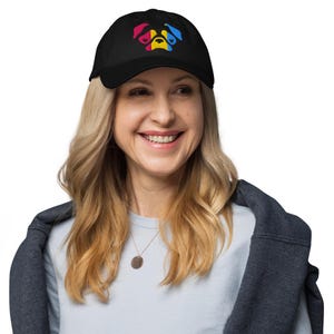 May include: Black baseball cap with a colorful paw print design. The paw print features red, yellow, and blue colors. The cap is worn by a person with blonde hair and a light blue shirt.