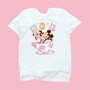 May include: White t-shirt with a cartoon image of Mickey Mouse and Minnie Mouse in a teacup.  The teacup has pink and white swirls.  There are three pastel colored lanterns above the teacup.