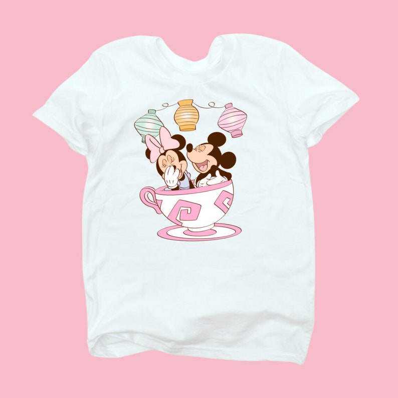 May include: White t-shirt with a cartoon image of Mickey Mouse and Minnie Mouse in a teacup.  The teacup has pink and white swirls.  There are three pastel colored lanterns above the teacup.
