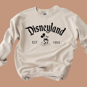 May include: A beige sweatshirt with a black graphic of Mickey Mouse and the text "Disneyland EST 1955".