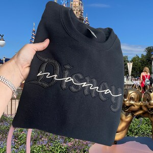 May include: Black sweatshirt with a white embroidered "Disney" logo. The word "mama" is written in cursive underneath the logo.