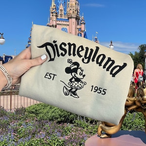 K&ouml;nnte beinhalten: Beiges Produkt mit schwarzem gesticktem Schriftzug "Disneyland", Minnie Mouse-Grafik und Text "EST 1955". Im Hintergrund ein Schloss und blauer Himmel.