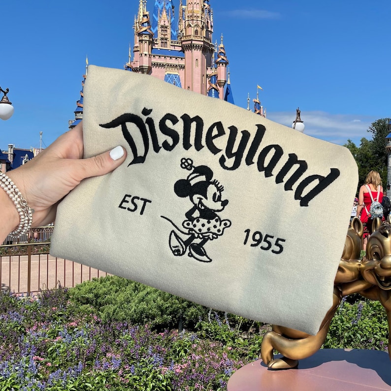 K&ouml;nnte beinhalten: Beiges Produkt mit schwarzem gesticktem Schriftzug "Disneyland", Minnie Mouse-Grafik und Text "EST 1955". Im Hintergrund ein Schloss und blauer Himmel.