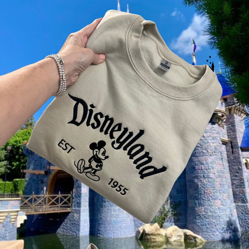 Puede incluir: Una sudadera beige con un logotipo de Disneyland negro, Mickey Mouse y el texto "EST 1955".