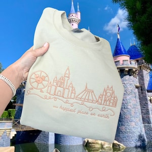 Puede incluir: Sudadera beige claro con un dise&ntilde;o bordado en color &oacute;xido de un horizonte de parque tem&aacute;tico. El dise&ntilde;o incluye una noria, un castillo y otras estructuras, con el texto "... happiest place on earth".