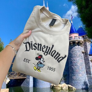 May include: A beige sweatshirt with a black embroidered Disneyland logo and a small embroidered image of Minnie Mouse. The text "EST 1955" is also embroidered on the sweatshirt.
