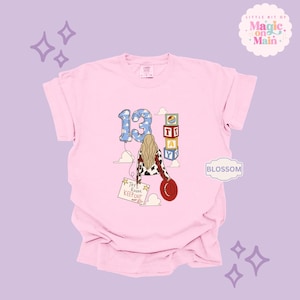 May include: A light pink t-shirt featuring a graphic of a person with long blonde hair wearing a cow-print jacket and holding a red hat. The design includes a blue cloud-patterned number 13 balloon, stacked alphabet blocks spelling TAY, and a sign reading Tay's Room Keep Out.