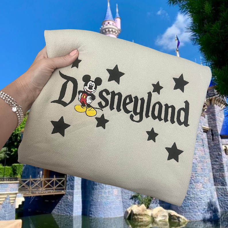 K&ouml;nnte beinhalten: Eine beige Tragetasche mit einem schwarzen gestickten Mickey Mouse und dem Text "Disneyland" umgeben von schwarzen Sternen.