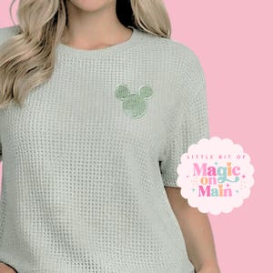 May include: A light sage green waffle-knit short-sleeve shirt featuring a small, embroidered mouse-ear silhouette patch on the left chest. A circular logo with a scalloped edge displays the text Little Bit of Magic on Main in pink and blue.