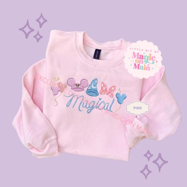 May include: A light pink sweatshirt with the word "Magical" in blue script, embroidered with cartoon character icons. The design includes a mouse head, a wizard hat, and a bow. The text "LITTLE BIT OF MAGIC ON MAIN" is on a white label.