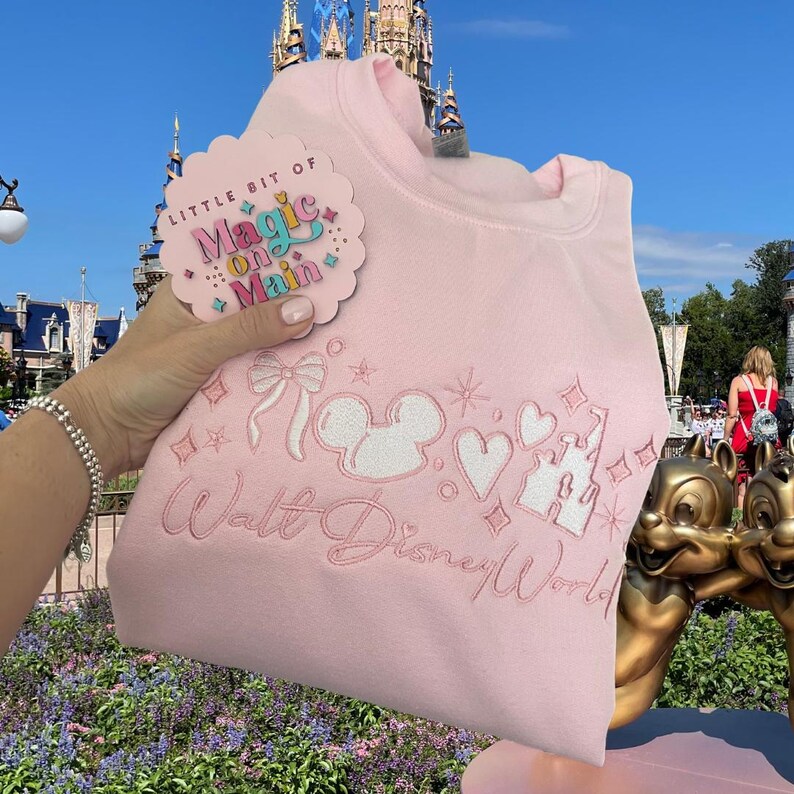 Może przedstawiać: R&oacute;żowa bluza z haftowaną białą głową Myszki Miki i napisem "Walt Disney World" oraz r&oacute;żową i białą przypinką z napisem "Little Bit of Magic on Main".