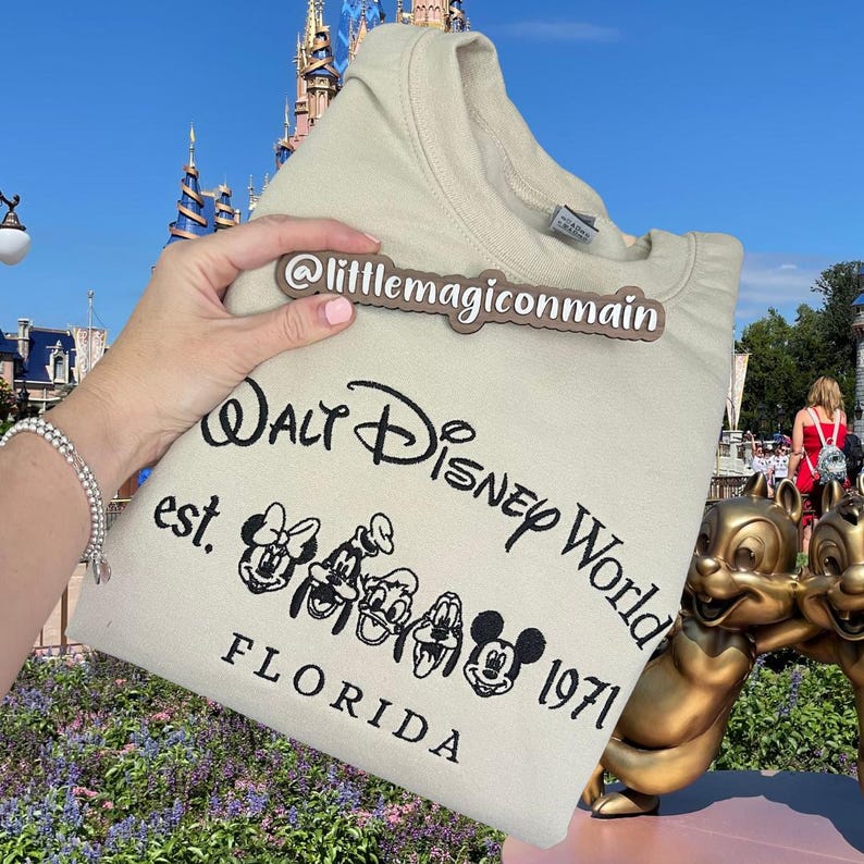 May include: A beige sweatshirt with a black embroidered design that says "Walt Disney World est. 1971 Florida" with Mickey Mouse and friends. The sweatshirt is being held by a hand with a brown tag that says "@littlemagiconnain".