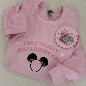 May include: Pink sweatshirt with the text "I am literally just a Disney girl" and a black embroidered Mickey Mouse ears headband. A small pink and white circle sticker with the text "Little bit of magic on Main" is attached to the sweatshirt.