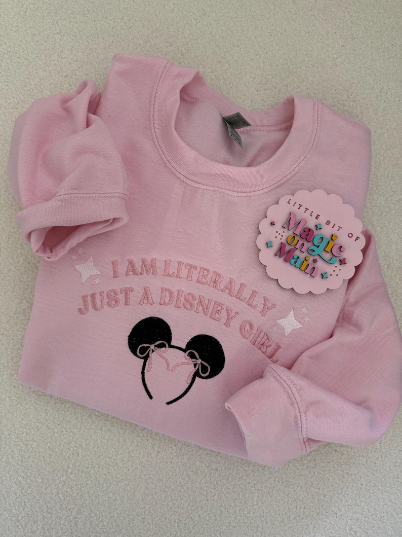 May include: Pink sweatshirt with the text "I am literally just a Disney girl" and a black embroidered Mickey Mouse ears headband. A small pink and white circle sticker with the text "Little bit of magic on Main" is attached to the sweatshirt.