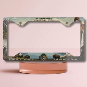 May include: License plate frame with a vintage National Parks design. The frame features illustrations of mountains, waterfalls, bison, and the text "EXPLORE, PROTECT, PRESERVE, ENJOY". The top reads "NATIONAL PARKS - AMERICA'S BEST IDEA".