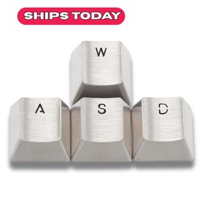 Könnte beinhalten: Nahaufnahme von vier silberfarbenen Tastenkappen mit den Buchstaben "W", "A", "S" und "D". Die Tasten sind in einer Gaming-Konfiguration angeordnet. Der Text "SHIPS TODAY" steht in einem rot-weißen Banner.