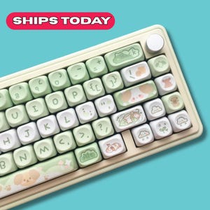 May include: A pastel-colored mechanical keyboard with a cream-colored frame and light green and white keycaps. The keycaps feature cute animal illustrations and decorative patterns. The keyboard has a round dial and a red banner that says "SHIPS TODAY".