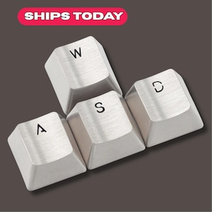Può includere: Quattro tasti di tastiera color argento, disposti in gruppo, con le lettere "W", "A", "S" e "D" stampate sopra. I tasti hanno una texture in metallo spazzolato. Il testo "SHIPS TODAY" è in un rettangolo rosa in alto a sinistra.