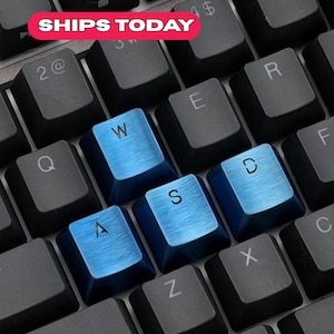 Puede incluir: Primer plano de un teclado de ordenador con teclas negras y teclas azules que deletrean "WASD". Las teclas tienen un aspecto de metal cepillado. El texto "SHIPS TODAY" está en una pancarta roja y blanca en la esquina superior izquierda.