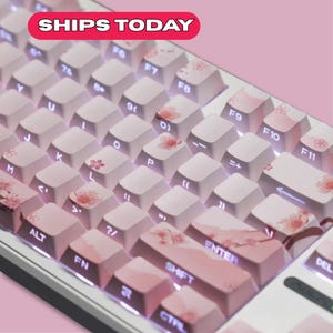 May include: A white and pink mechanical keyboard with cherry blossom keycaps. The keycaps have a light pink base with cherry blossom designs. The keyboard has a white frame and the text "SHIPS TODAY" in a red banner.