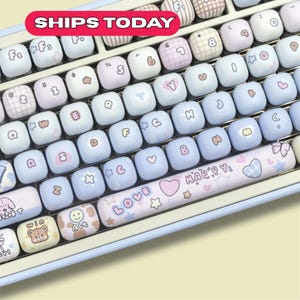 Może przedstawiać: Klawiatura w pastelowych kolorach z niestandardowymi nakładkami na klawisze. Klawisze są jasnoniebieskie, białe i różowe, ozdobione sercami, gwiazdkami i innymi uroczymi wzorami. Spacja ma napis "LOVE HAPPY". Klawiatura ma jasnoniebieską ramkę.