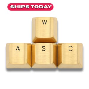 Può includere: Quattro tasti dorati per tastiera disposti a grappolo. I tasti sono contrassegnati con le lettere "W", "A", "S" e "D". Il testo "SHIPS TODAY" è in uno striscione rosso e bianco in alto.