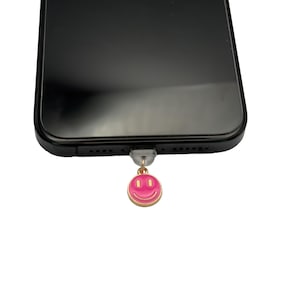 Könnte beinhalten: Ein rosa Smiley-Anhänger mit goldener Umrandung hängt an einem schwarzen Smartphone.