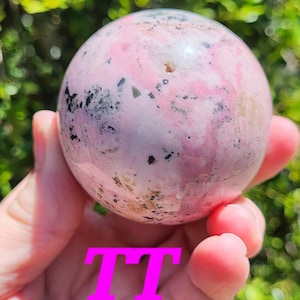 K&ouml;nnte beinhalten: Eine rosa-graue Kugel mit einem gesprenkelten Muster wird in einer Hand gehalten. Die Kugel hat eine polierte Oberfl&auml;che und scheint ein Edelstein oder ein dekoratives Objekt zu sein. Die Buchstaben "TT" sind auf dem Bild &uuml;berlagert.