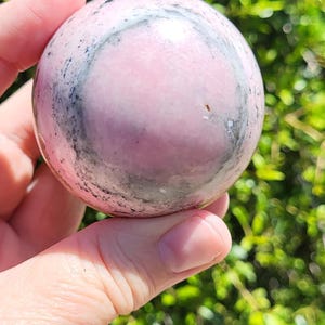 K&ouml;nnte beinhalten: Eine rosa Kugel mit grau-schwarzer Marmorierung wird in einer Hand vor einem Hintergrund aus gr&uuml;nem Laub gehalten. Die Kugel ist glatt und poliert. Die Buchstaben "TT" befinden sich in der unteren rechten Ecke.