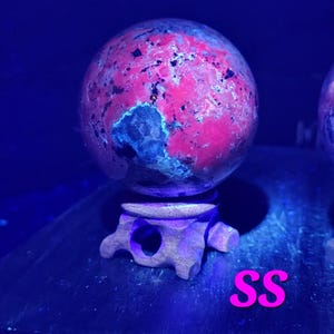 K&ouml;nnte beinhalten: Eine rote und schwarze Steinkugel auf einem Holzst&auml;nder. Die Kugel hat ein gesprenkeltes Aussehen mit dunklen Flecken. Der St&auml;nder ist aus dunklem Holz mit einem geschnitzten Design. Die Buchstaben "SS" sind rosa.