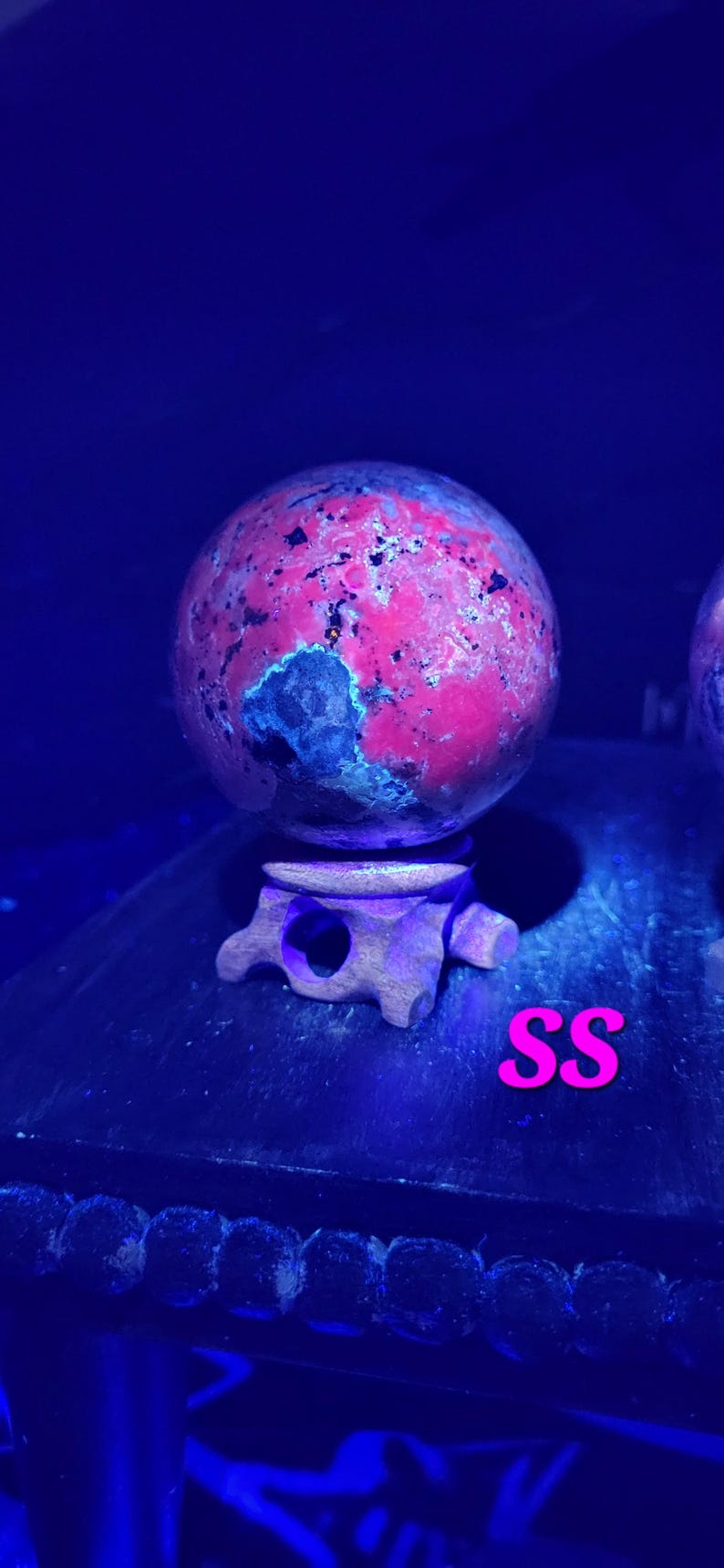 K&ouml;nnte beinhalten: Eine rote und schwarze Steinkugel auf einem Holzst&auml;nder. Die Kugel hat ein gesprenkeltes Aussehen mit dunklen Flecken. Der St&auml;nder ist aus dunklem Holz mit einem geschnitzten Design. Die Buchstaben "SS" sind rosa.