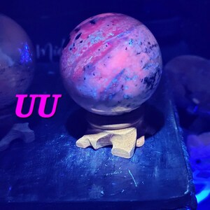 K&ouml;nnte beinhalten: Ein kugelf&ouml;rmiger, polierter Stein mit einem wirbelnden Muster aus rosa, lila und blauen Farbt&ouml;nen, montiert auf einem Holzst&auml;nder. Der Stein wird von blauem Licht beleuchtet, und die Buchstaben "UU" sind im Vordergrund sichtbar.