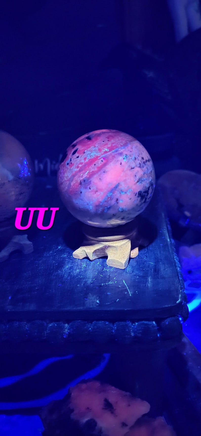 K&ouml;nnte beinhalten: Ein kugelf&ouml;rmiger, polierter Stein mit einem wirbelnden Muster aus rosa, lila und blauen Farbt&ouml;nen, montiert auf einem Holzst&auml;nder. Der Stein wird von blauem Licht beleuchtet, und die Buchstaben "UU" sind im Vordergrund sichtbar.