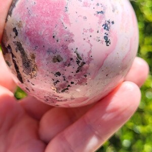 K&ouml;nnte beinhalten: Eine rosa und wei&szlig;e Kugel mit dunkelgrauen und schwarzen Markierungen. Die Kugel wird in einer Hand gehalten, mit einem verschwommenen gr&uuml;nen Hintergrund aus Bl&auml;ttern. Die Kugel ist wahrscheinlich ein Dekorationsartikel oder ein Kristall.
