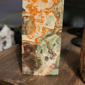 May include: A green, orange, and white polished stone with a pointed top. The stone is shaped like a pyramid and has a natural, organic pattern.