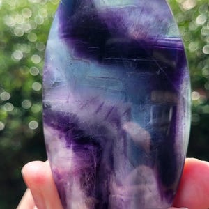 May include: A polished, teardrop-shaped fluorite crystal. The stone displays a blend of purple, blue, and green hues, with visible internal patterns. The crystal is held against a blurred green background.
