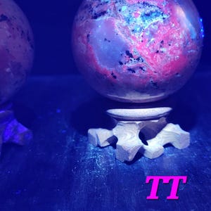 K&ouml;nnte beinhalten: Zwei kugelf&ouml;rmige Dekorationsgegenst&auml;nde mit einem rosa und blauen Muster, die auf einem Holzst&auml;nder ruhen. Die Objekte werden von blauem Licht beleuchtet, und die Buchstaben "TT" sind in Pink zu sehen.