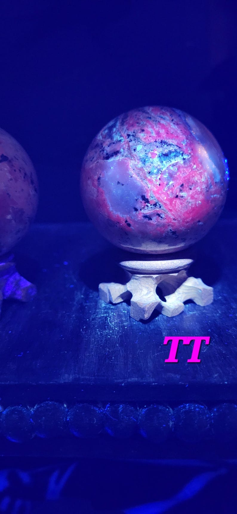 K&ouml;nnte beinhalten: Zwei kugelf&ouml;rmige Dekorationsgegenst&auml;nde mit einem rosa und blauen Muster, die auf einem Holzst&auml;nder ruhen. Die Objekte werden von blauem Licht beleuchtet, und die Buchstaben "TT" sind in Pink zu sehen.