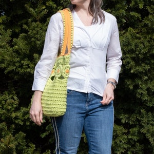 Pu&ograve; includere: Una donna indossa una camicia bianca abbottonata e jeans blu. Porta una borsa a mano in crochet verde con accenti arancioni e gialli.