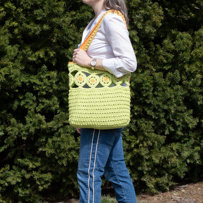 Pu&ograve; includere: Una borsa tote in crochet verde lime con bordi arancioni e un design floreale. La borsa ha una base larga e piatta e un manico superiore.