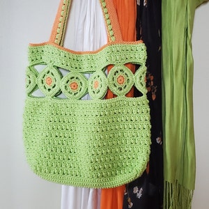 Pu&ograve; includere: Una borsa a mano in crochet verde con dettagli arancioni appesa a un appendiabiti con altre sciarpe. La borsa ha un design unico con cerchi aperti e un motivo testurizzato.