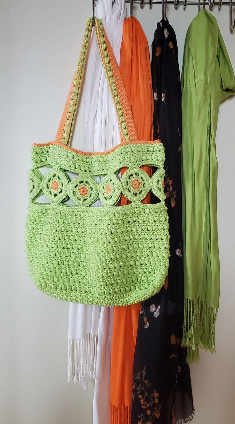 Pu&ograve; includere: Una borsa a mano in crochet verde con dettagli arancioni appesa a un appendiabiti con altre sciarpe. La borsa ha un design unico con cerchi aperti e un motivo testurizzato.